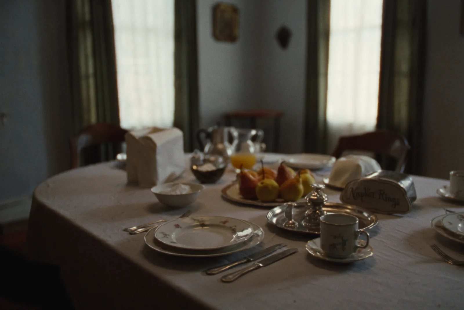 A breakfast table set for four with a white cloth, mismatched antique china, a glass pitcher of juice, a coffee pot and a bowl of pears, with tall curtained windows behind.