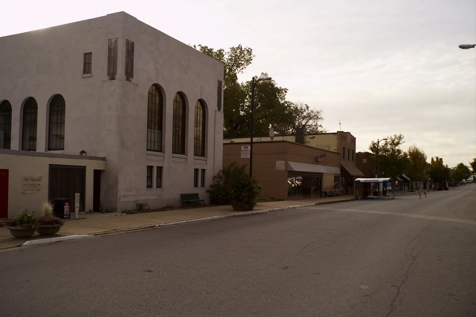 A downtown street with a grand limestone civic building with tall arched windows on one side, low brick storefronts opposite, a wide sidewalk with planters and a wooded hill rising behind the rooftops.