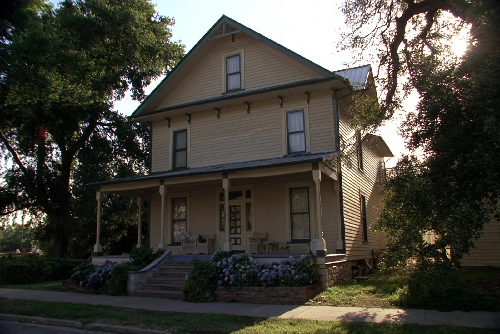 A restored two-story clapboard house painted soft cream with dark green trim, a deep porch with turned posts and wicker chairs, stone steps up from the sidewalk and mature oaks in the late afternoon sun.