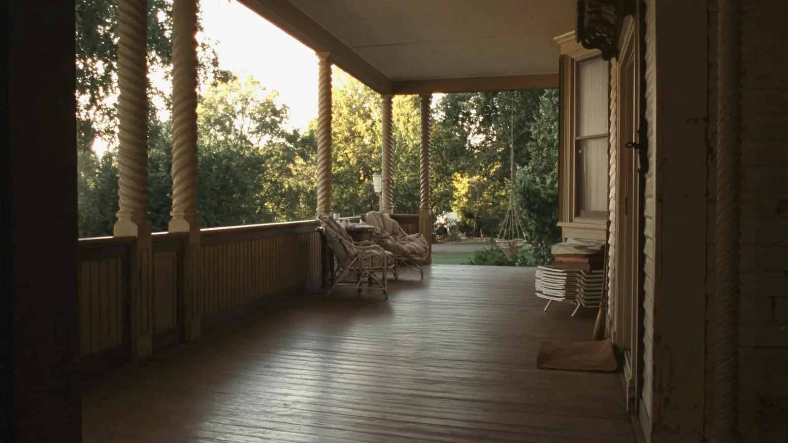 The deep front porch of an old clapboard house seen from inside, with turned posts, painted grey floorboards, two wicker chairs with faded cushions, a small table between them and a swing at the far end.
