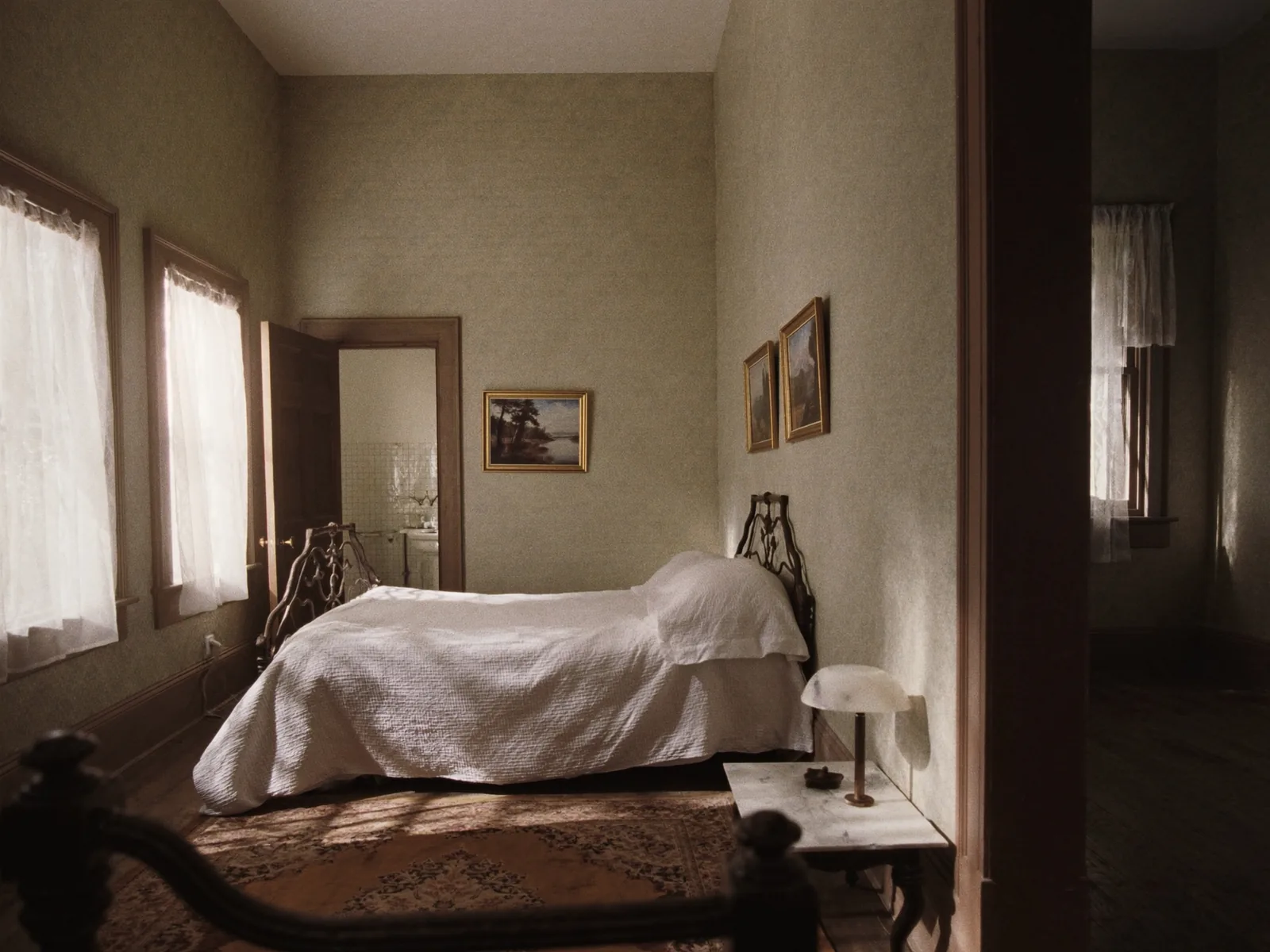 A guest bedroom with an antique iron bed under a white quilt, a marble-topped nightstand with a lamp, muted sage patterned wallpaper, a worn rug on a wooden floor and an open door showing a tiled bathroom.